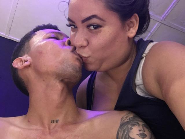 ElChicletv with a shaved head and tattoo on the right shoulder, kissing another ElChicletv with dark hair and a tank top, both puckering up. Purple light in the background.