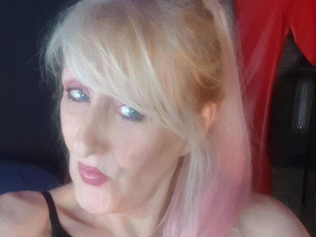 LadyHazel, con cabello rubio, puntas rosas, ojos azules y lápiz labial rosa, vestida con un top negro, mirando ligeramente hacia un lado, con un paño rojo en el fondo..