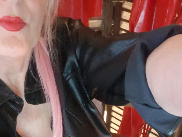 LadyHazel, con cabello rosa, chaqueta de cuero negro y lápiz labial rojo, posando en un selfie mostrando su escote y hombro descubierto. Los senos son parcialmente visibles. La parte superior del cuerpo y el brazo están enfocados..