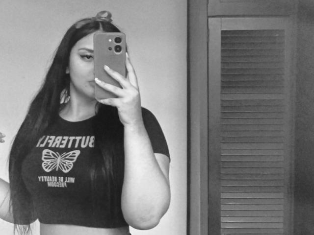AmaliaDalton with long dark hair, holding a phone, wearing a butterfly crop top, showing her tits, in a black-and-white selfie, with a wooden shutter in the background.