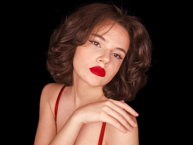 JunneWers hat kurze, braune, lockige Haare, roten Lippenstift und rote Träger ihres Kleides. Ihre Hand berührt ihren Hals, und im Hintergrund ist Schwarz zu sehen. Ihre Vulva ist nicht sichtbar..