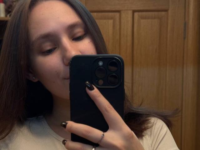 HotCaramells with long brown hair, fair skin, and black nails, taking a selfie with a black phone, wearing a white shirt, and a nose ring.