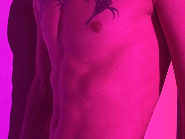 NikkyBanny's bare chest with a small tattoo above the nipple, pink-purple lit skin, visible ribs, and a slight shadow on the left side. NikkyBanny's flat, toned stomach.