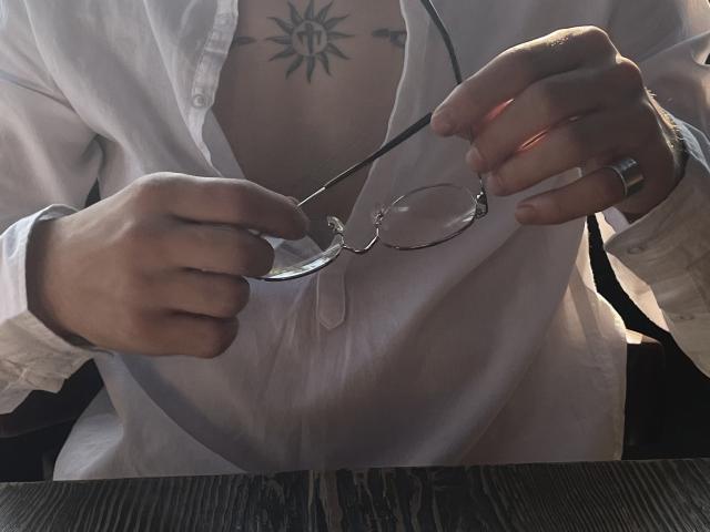 NikkyBanny's hands hold round glasses with a black frame, wearing a white shirt, exposing a sun tattoo on their chest, with a silver ring on their right hand.