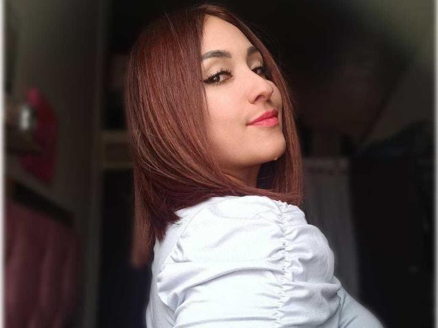 ImYourSophie has straight brown hair, light skin, and is wearing a white shirt with ruffled sleeves, looking back with a slight smile and pink lipstick.