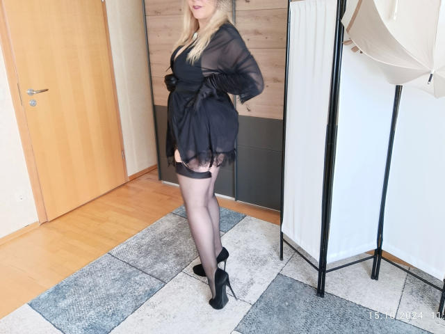 Rassige-Frau with blonde hair, black sheer dress, black garter, black stockings, and high heels, standing in a room, showing her ass, in a sexy pose.