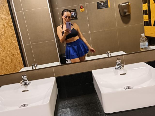 GalaMalkova in a bathroom selfie, wearing a black sports bra and blue skirt, showing her tits and ass in the mirror, holding a phone, with two white sinks.