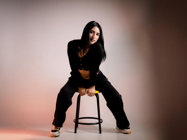 NickiClark is sitting on a stool with her legs spread, wearing a black top and pants, showing her tits and midriff, with a playful smile and long black hair.