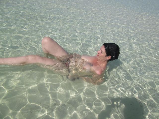 Sexy-Noemy-hot lying in clear water, legs spread, hands covering her pussy, water rippling over her tits, smiling, with dark wet hair, light skin, and a relaxed posture.