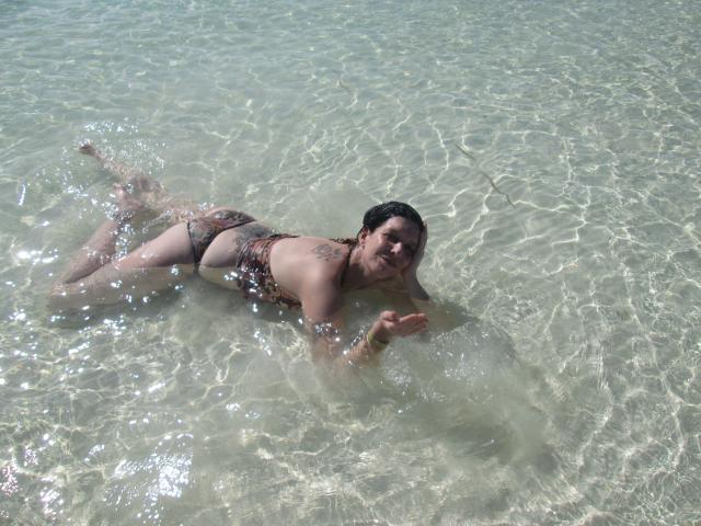 Sexy-Noemy-hot, lying face-down in clear water, wearing a leopard-print bikini, showing her ass and tattoo on her left side, with wet dark hair and a relaxed expression.