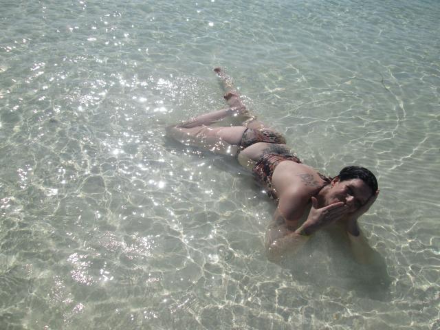 Sexy-Noemy-hot lies in clear water, wearing a bikini, with tattoos on her back and arm, legs together, hands near her face, sunlight sparkling on the water's surface.