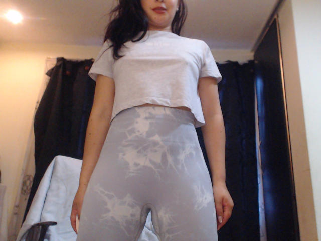 WendySchulz has light gray crop top and high-waisted, tie-dye, gray leggings, standing with arms at sides, black curtains in background, subtle smile, dark wavy hair.
