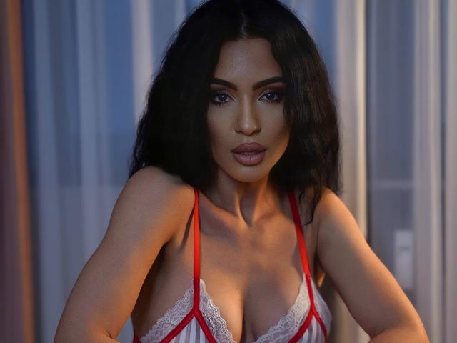 Aleciax with long black hair, wearing a red and white lace lingerie top, showing her tits, looking directly at the camera with a serious expression.