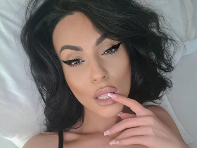 Aleciax, con i capelli neri ondulati, sopracciglia folte, eyeliner nero intenso, labbra rosa lucide e un dito che tocca le sue labbra. Ha la pelle abbronzata ed è sdraiata su cuscini bianchi..