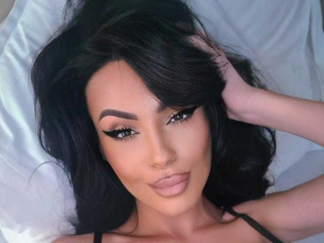 Aleciax has dark, wavy hair, thick eyebrows, long eyelashes, and full lips with nude lipstick. She's lying on a white bed, hand in hair, smiling subtly.