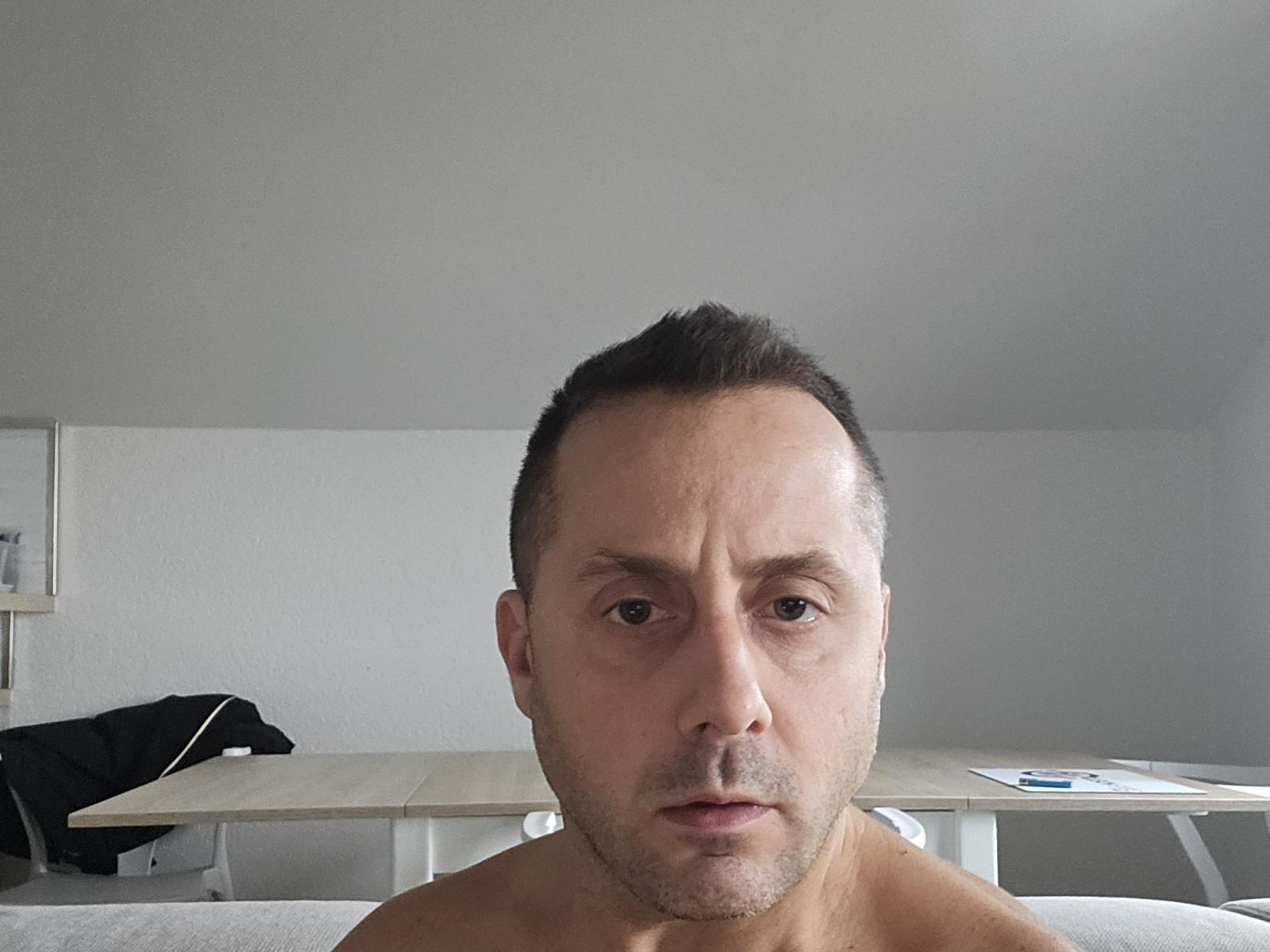 FrankyLeMagnifique's face is in the center, shirtless with a neutral expression, short dark hair, and a slightly balding forehead, in a plain room with a wooden table.
