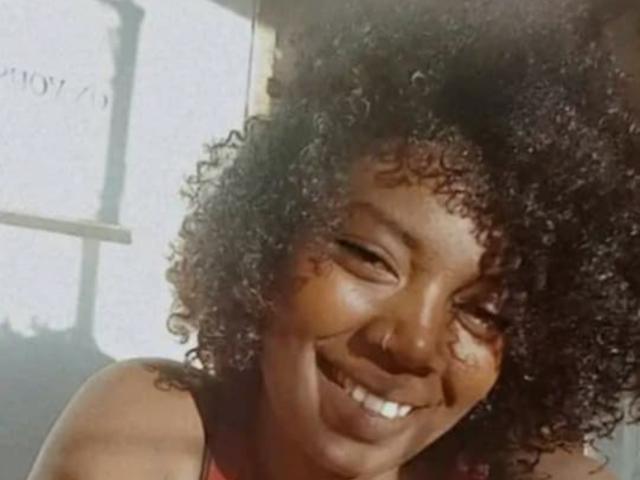 JessicaBella69 with dark curly hair, smiling with her eyes closed, showing her teeth, wearing a red sleeveless top, her brown skin glistening in the sunlight.