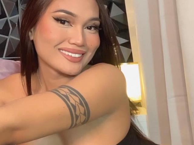 MissReign with brown hair, smiling, showing tattoo on her right arm, has bare shoulders, and is wearing eyeliner and pink lipstick, standing near a lit lamp.