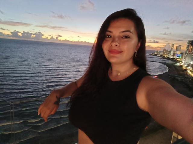 KatherinLatin with long dark hair, wearing a black crop top, showing her boobs, standing by a waterfront, sunset in the background, smiling at the camera.