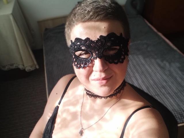 RachelExcite with short brown hair, wearing a black lace mask and choker, smiling at the camera, showing her small tits and a cross necklace, sitting on a bed.
