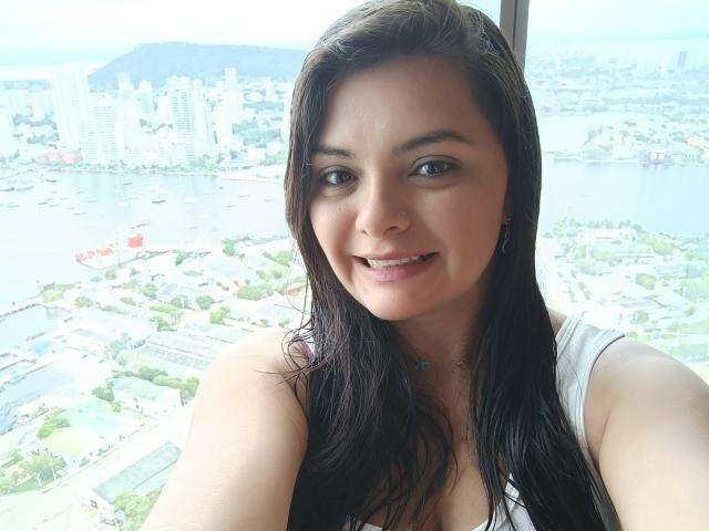 KatherinLatin has wet brown hair, small tits, and a cute smile, wearing a white sleeveless top, with a city and water view in the background.