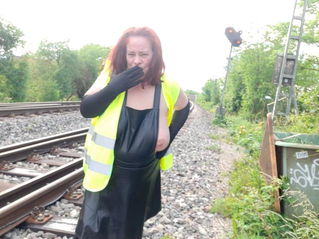 Kathy75-hot with red hair, wearing a neon yellow vest and black latex dress, black gloves, covering her mouth, standing on railroad tracks with gravel and greenery.