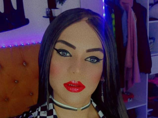 GeishaSenier has glossy red lips, blue eyes with thick black eyeliner, dark brown straight hair, and a white choker. She's looking at the camera with a serious expression.