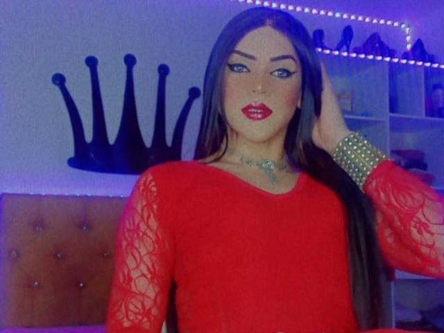 GeishaSenier with long brown hair, red lace long-sleeve top, red lipstick, green bracelet, and tattooed neck, hand in hair, standing in blue-lit room with crown wall decal.