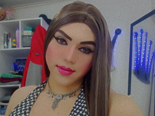 GeishaSenier has brown hair, thick eyebrows, pink lipstick, and a necklace with a floral design. She's wearing a black and white patterned top. Her tits are visible.
