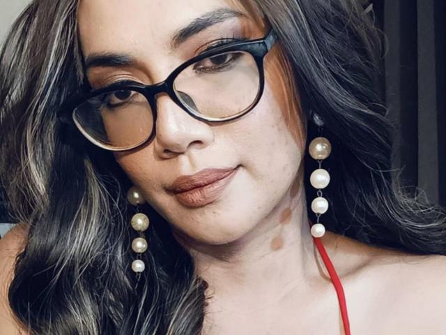 TransKathie with dark wavy hair, black glasses, pearl earrings, red halter, and brown lipstick, giving a subtle smirk. Her face has a few dark spots.