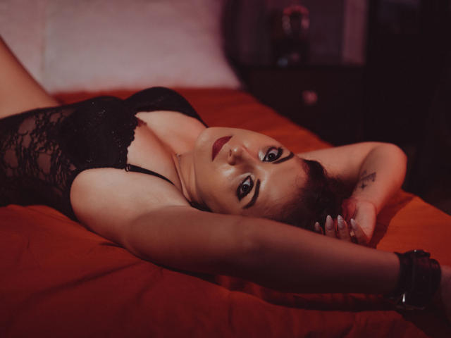 MistressCarla lying on an orange bed, wearing black lace lingerie, arms above her head, red lipstick, black wristband, looking at the camera, seductive, dark room, sexy.