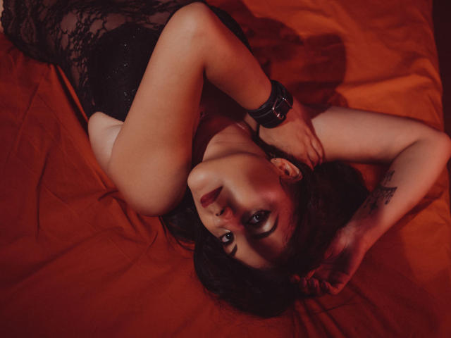 MistressCarla lying on red sheets, wearing black lace lingerie, with arms above head, black bracelet on wrist, looking up, dark hair, tattoo on right wrist, red light.