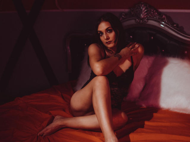 MistressCarla with dark hair, black lace lingerie, sitting on an orange bed, legs crossed, arms up, showing her tits and ass, looking at the camera in dim light.
