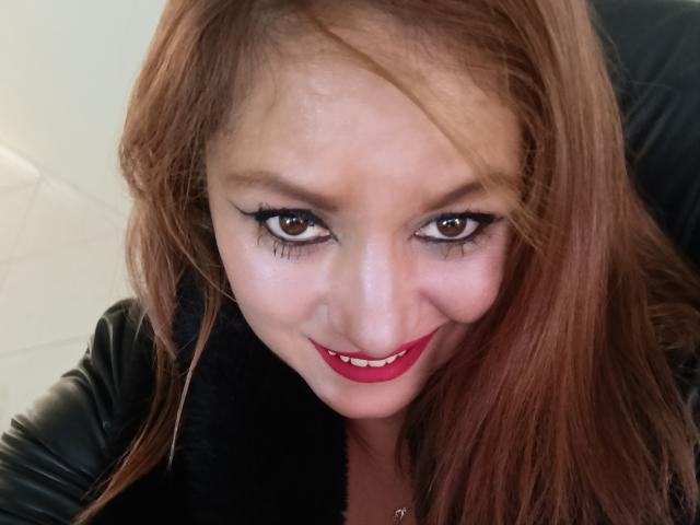 NatashaChaus with red hair, black leather jacket, and red lipstick, smiling at the camera with brown eyes and black eyeliner. Her tits are not visible.