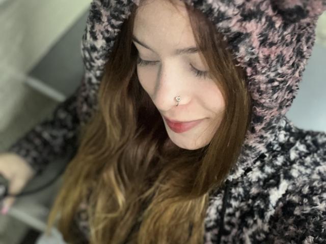 ShantalSummer with long brown hair, pink nose ring, and red lips, wearing a fluffy black-and-pink hoodie, holding a black object, looking down with a slight smile.
