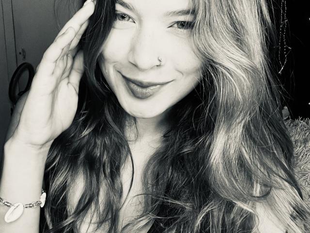 ShantalSummer has wavy, medium-length hair, a nose ring, and a subtle smile. She's touching her hair with her right hand, wearing a bracelet with a heart charm.