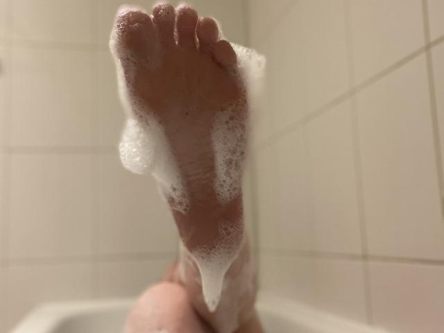 LexyClaire's soapy foot, with bubbles clinging to her toes, is the focus. Her leg is visible, and she's in a white-tiled bathtub. The shot is low and up-close.