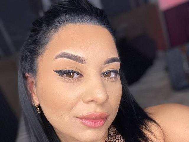 MadyLux hat eine gebräunte Haut, schwarzes Haar, dicke Augenbrauen, einen Eyeliner im "Winged"-Stil, pinken Lippenstift und ein leichtes Lächeln. Sie trägt ein Netzoberteil und kleine Ohrringe..
