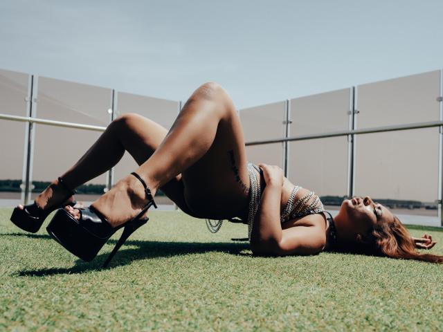 MadyLux est allongée sur l'herbe, en talons hauts noirs, portant un haut scintillant, avec les jambes levées et les fesses en l'air, sous un ciel bleu clair..