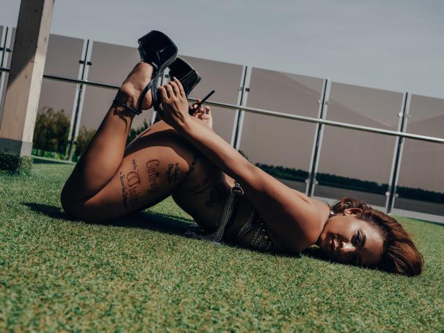 MadyLux liegt auf dem Rasen, auf dem Rücken, mit hochgestrecken Beinen, hält schwarze High Heels, trägt einen silbernen Bikini und zeigt Tattoos an ihren Oberschenkeln. Sie hat braunes Haar und ein verführerisches Lächeln..