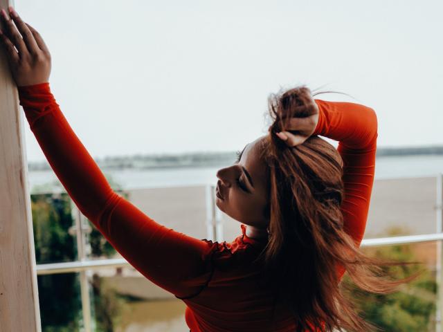 MadyLux mit langen, roten Haaren, die ein rotes, langärmeliges Oberteil trägt, hebt ihre Arme und hält ihr Haar hoch, wobei ihre Brüste sichtbar sind, und blickt mit einem ruhigen Ausdruck zur Seite..