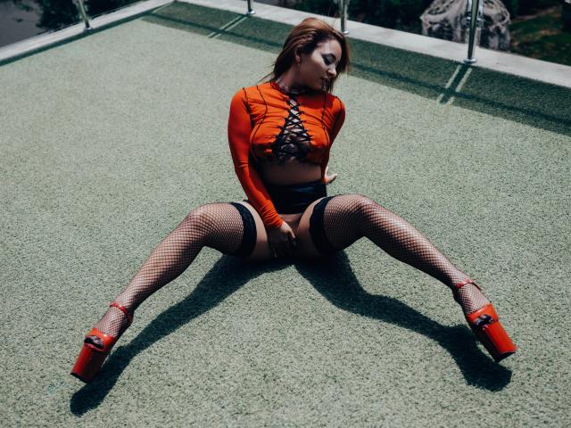 MadyLux con top arancione, lacci neri e tacchi alti rossi, seduta sull'erba verde, gambe divaricate, indossa calze a rete, mostrando il sedere e la vulva, con lo sguardo rivolto verso il basso..