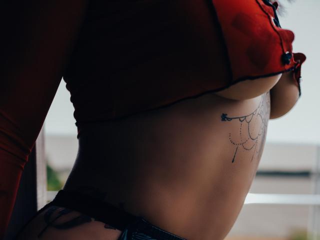 MadyLux indossa una parte superiore in rete rossa che mette in mostra i suoi seni e un tatuaggio delicato sotto il seno destro. Indossa delle mutandine in pizzo nero, con il ventre e il tatuaggio visibili..