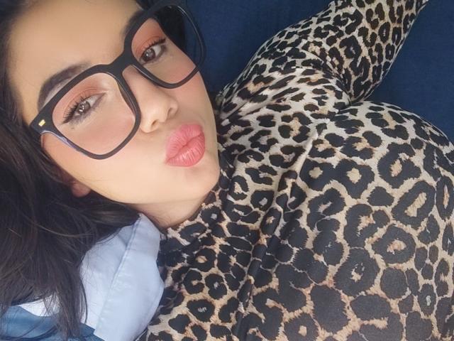 SquirtinGamy met volmaakte, roze lippen, gekleed in een luipaardprint body, zwarte bril en lang donker haar, liggend, met grote, ronde borsten..