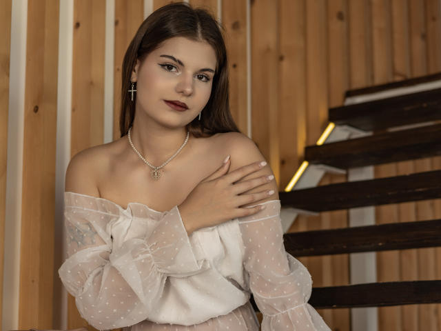 AdeliaLove with dark brown hair, wearing a sheer white polka dot top, exposing her shoulders and small tits, hands on chest, cross earrings, and a pearl necklace, standing by stairs.