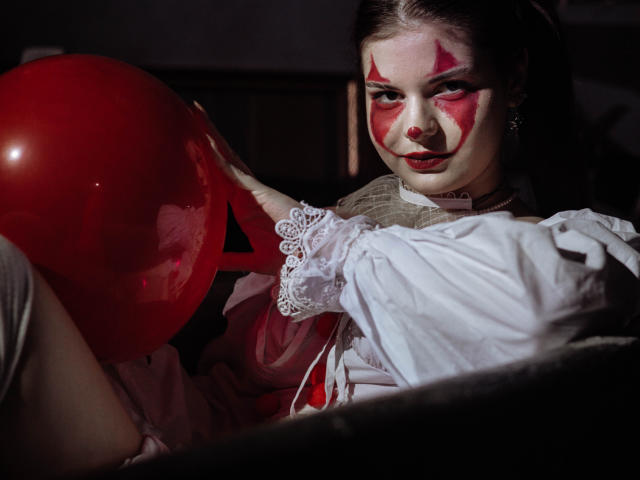 AdeliaLove with red face paint, white lace dress, holding a red balloon, dark eyes, slight smile, dark background, light on her face, and a pearl necklace.