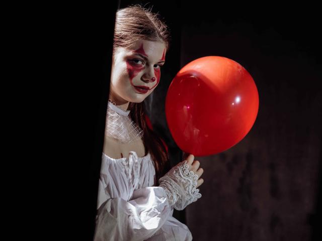 AdeliaLove with red makeup, white dress, and lace gloves, holding a red balloon, peeks from dark shadows, looking playful, her tits slightly visible under the dress.