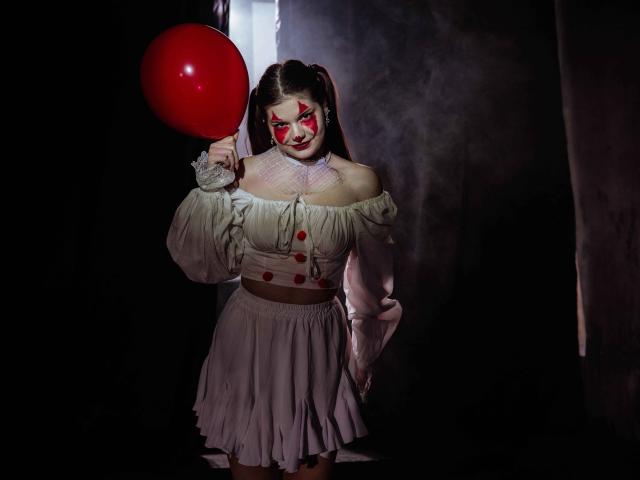 AdeliaLove with painted red clown face, holding red balloon, wearing off-shoulder white dress, puffy sleeves, and brown belt, standing in dark, smoky, dimly-lit space.