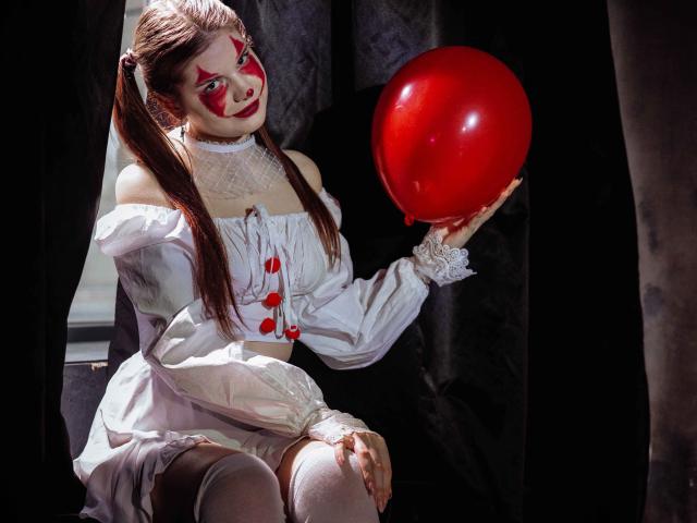 AdeliaLove with red balloon, wearing white off-shoulder dress, red face paint, and pigtails, sitting on black curtain, holding red balloon, showing white thigh-highs and lace cuffs.