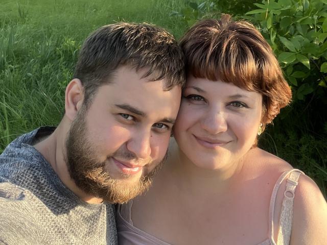 AlisaNAleks has short brown hair and a smile, standing next to a bearded dude with a gray hoodie, both with cute faces and soft skin, in a sunny grassy area.